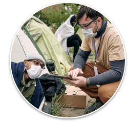 A Black man wearing a mask leans out of the tent he is staying in to talk to a health care worker, a white man who is also wearing a mask and is crouched down with a clipboard.