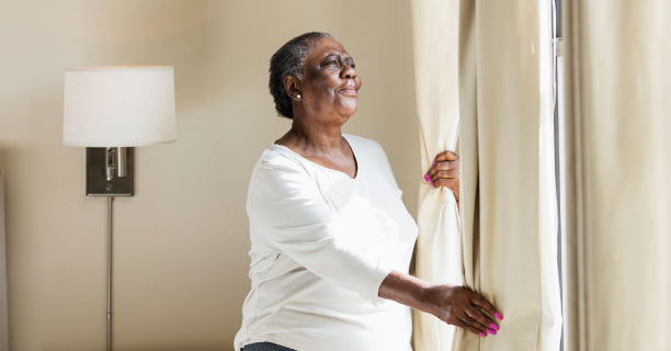 An older Black woman opening her curtain and looking out a sunny window.